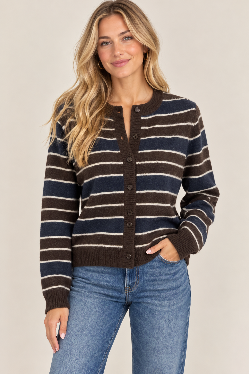 A woman modeling a brown, navy, and beige striped button-up cardigan sweater with blue jeans, set against a plain beige studio background.