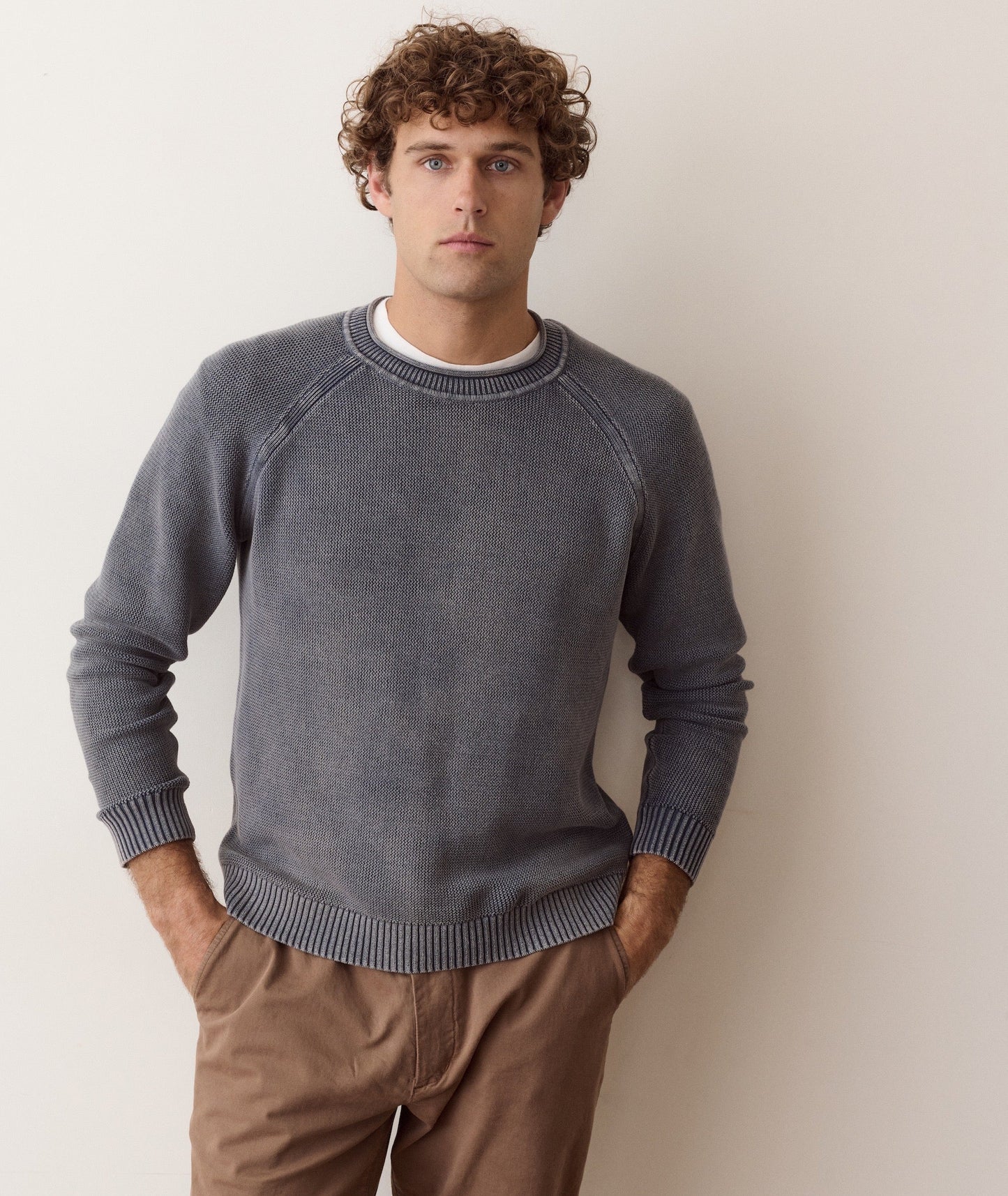A man wearing a dark gray knit crewneck sweater with raglan sleeves, paired with brown pants, standing against a plain light beige wall.