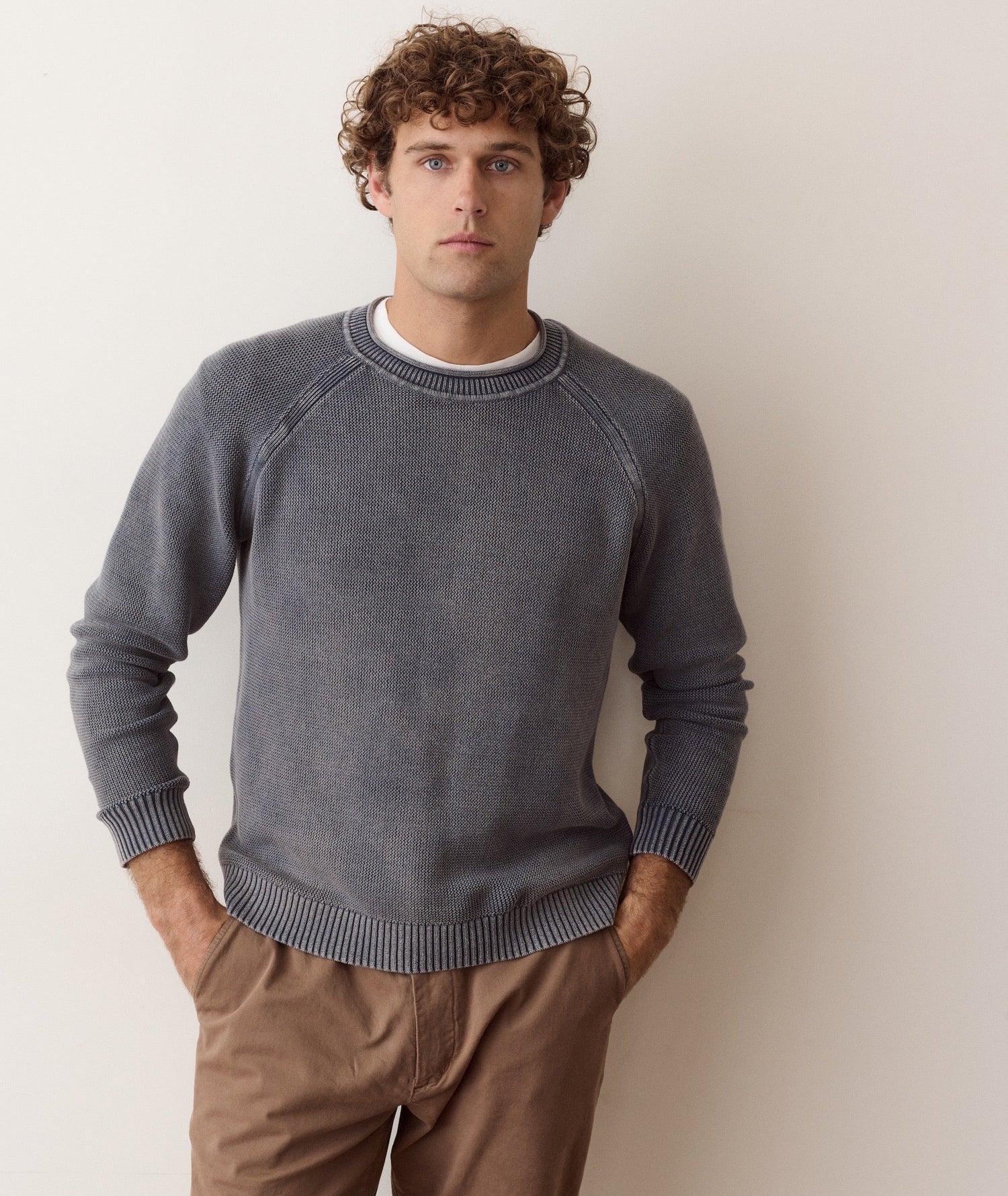 A man wearing a dark gray knit crewneck sweater with raglan sleeves, paired with brown pants, standing against a plain light beige wall.