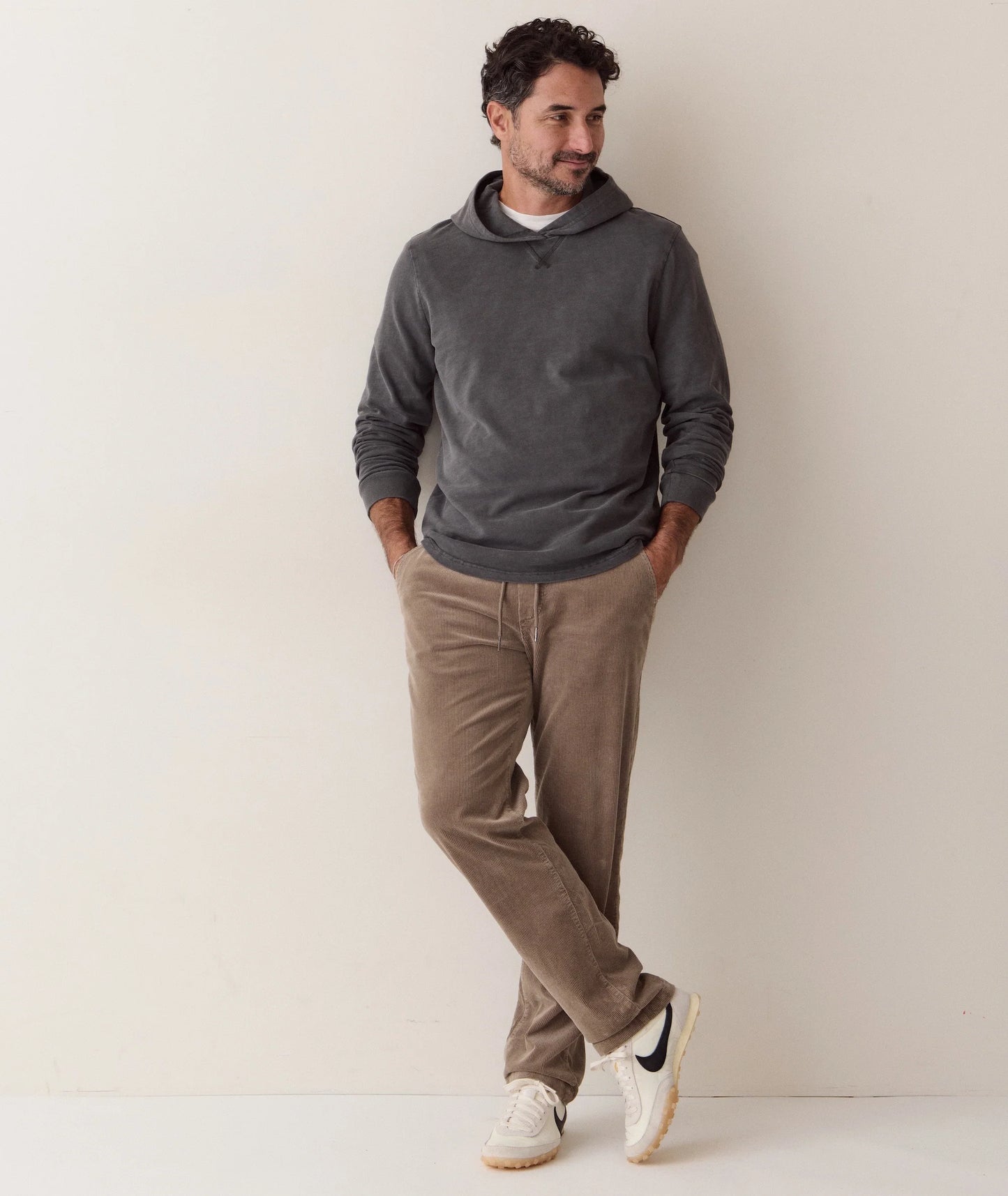 A man wearing a dark gray long-sleeve hoodie, brown corduroy pants, and white sneakers, leaning against a plain off-white wall.