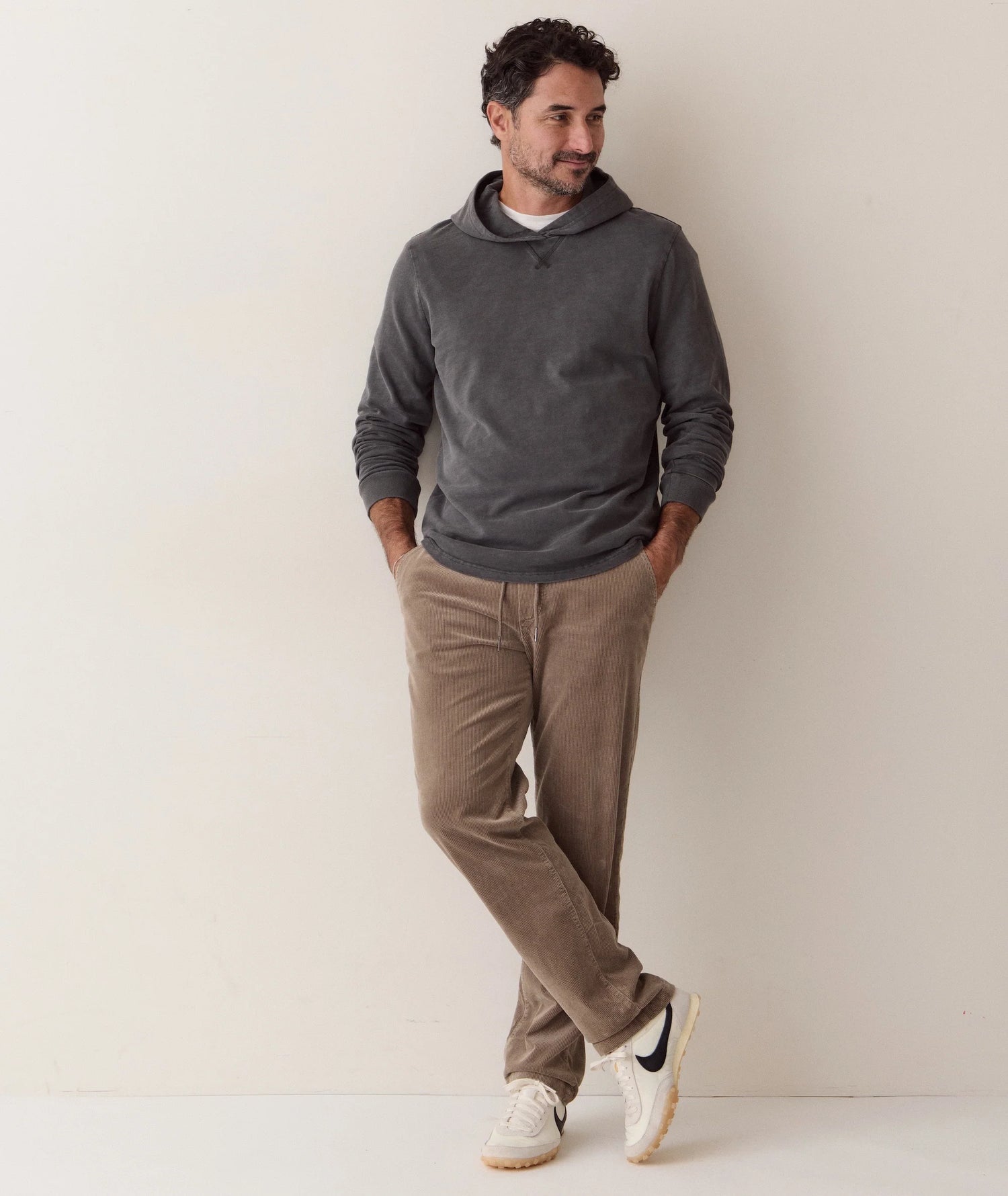 A man wearing a dark gray long-sleeve hoodie, brown corduroy pants, and white sneakers, leaning against a plain off-white wall.