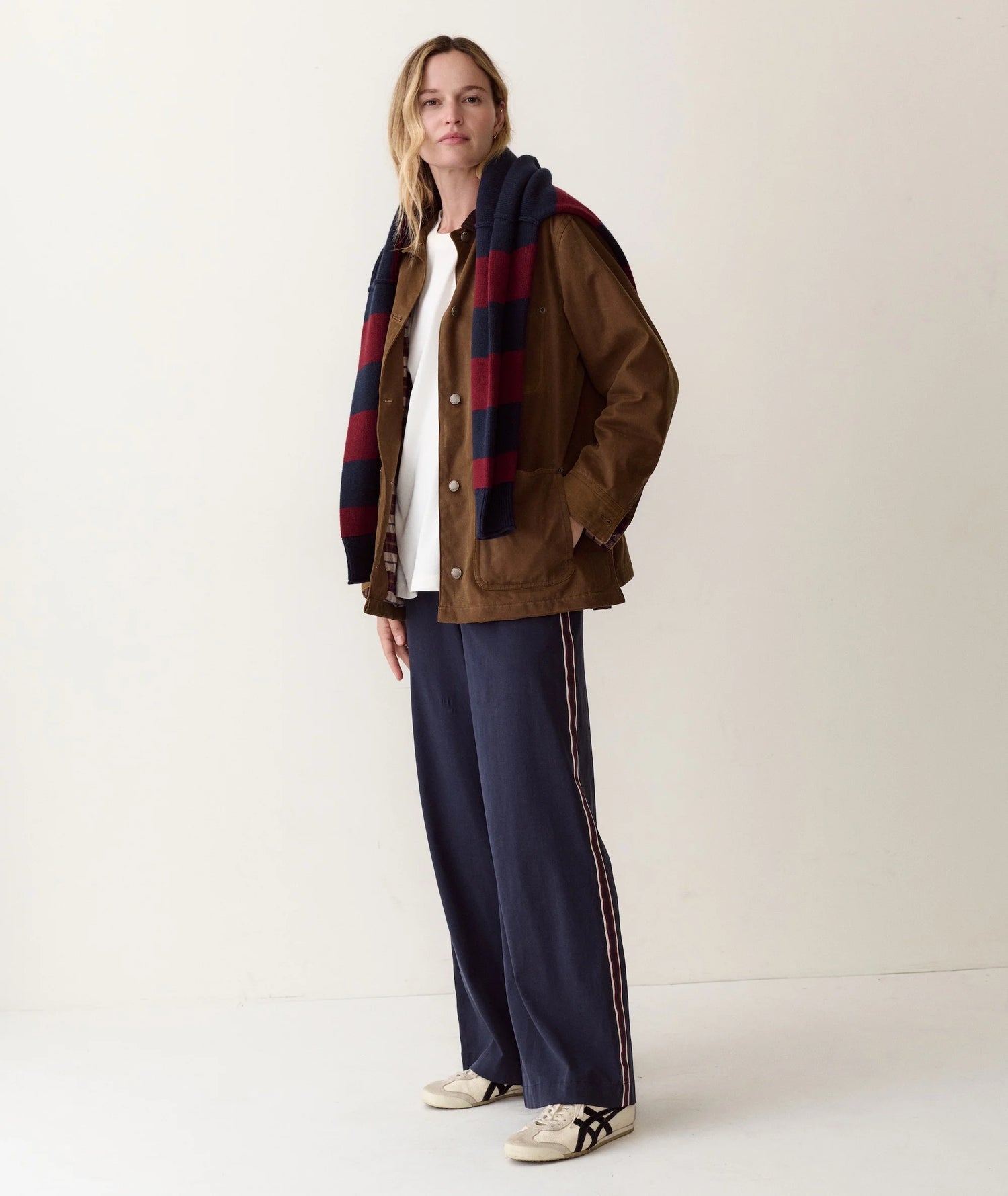 A woman stands against a plain off-white studio background wearing a brown jacket over a white top, a navy and red plaid scarf, navy wide-leg pants with thin light stripes, and white sneakers.